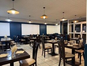 A restaurant with multiple tables and chairs, ceiling lights, and a large window. - Hotel De La Poste (Espinasses)