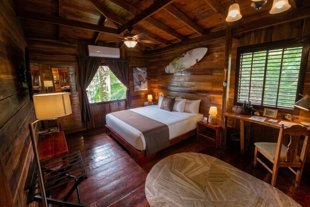 A wooden cabin-style room with a bed, desk, chair, and a window with blinds.