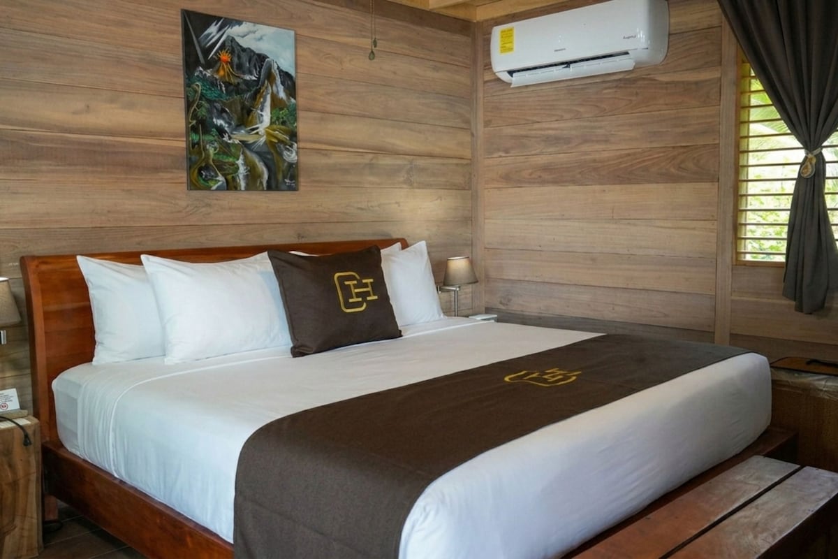 A hotel room with a wooden headboard, a bed with white bedding and a brown throw, an air conditioning unit, a nightstand with a lamp, and a window with curtains.
