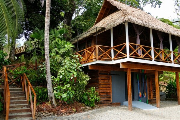 A wooden building with a thatched roof, surrounded by lush greenery and a wooden staircase leading to a balcony.