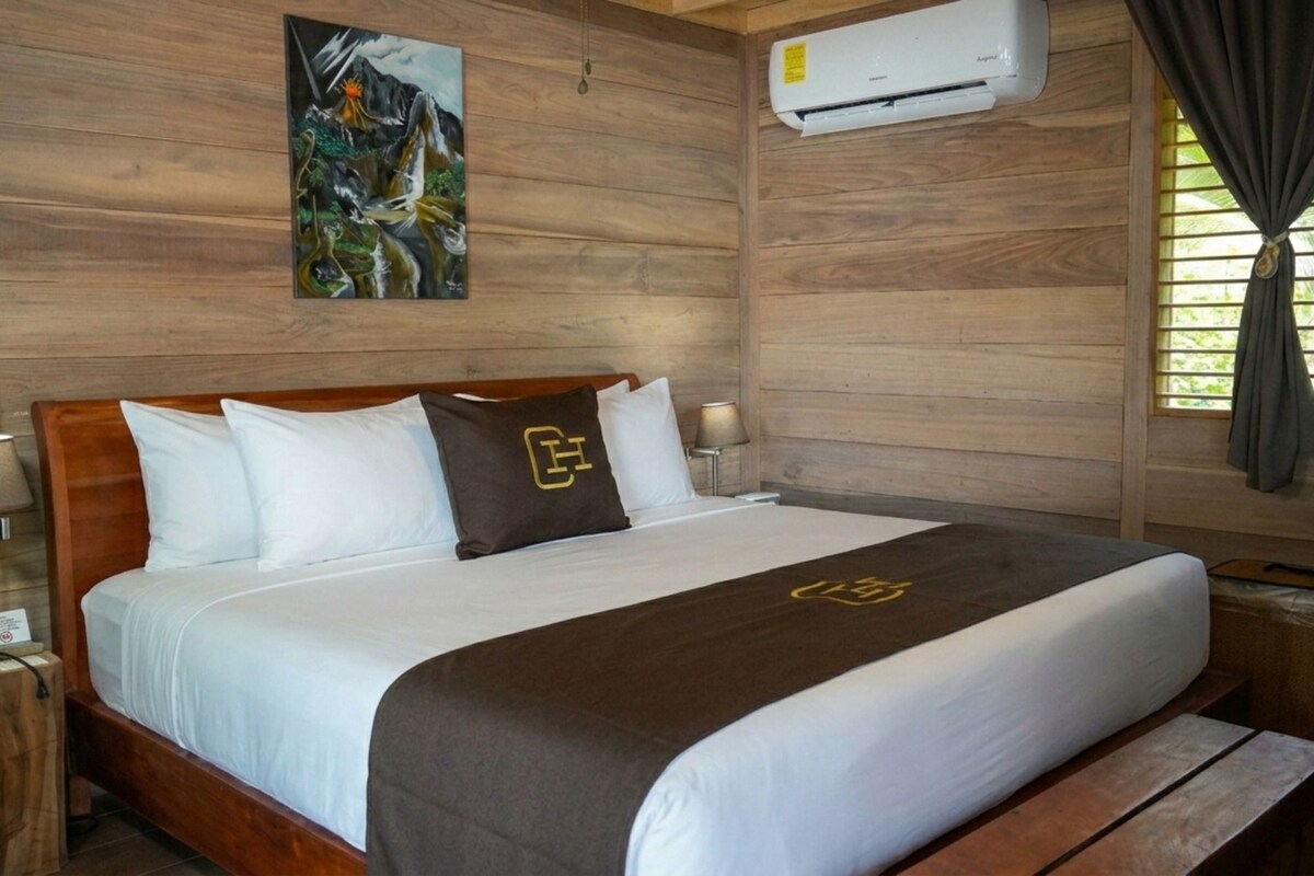 A hotel room with a wooden headboard, a bed with white bedding and a brown throw, an air conditioning unit, a nightstand with a lamp, and a window with curtains.