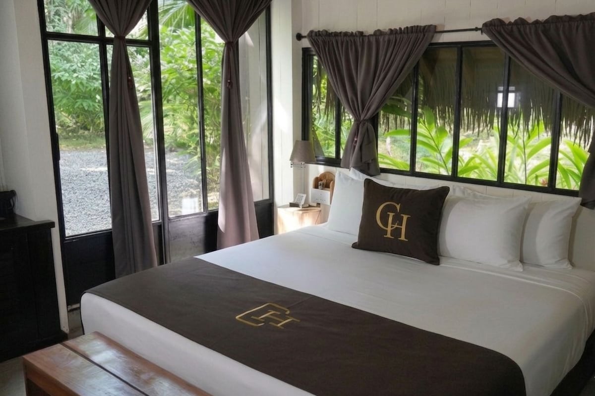 A hotel room with a bed, curtains, a bedside table with a lamp, and a view of greenery outside.