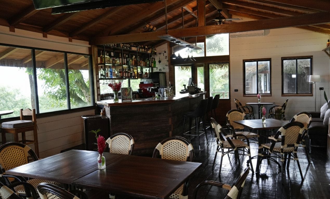A spacious indoor area with a bar, wooden tables, wicker chairs, and large windows offering a view of greenery outside.