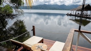 Una terraza de madera con una mesa y dos sillones de relax con vistas a un lago tranquilo y montañas al fondo.
