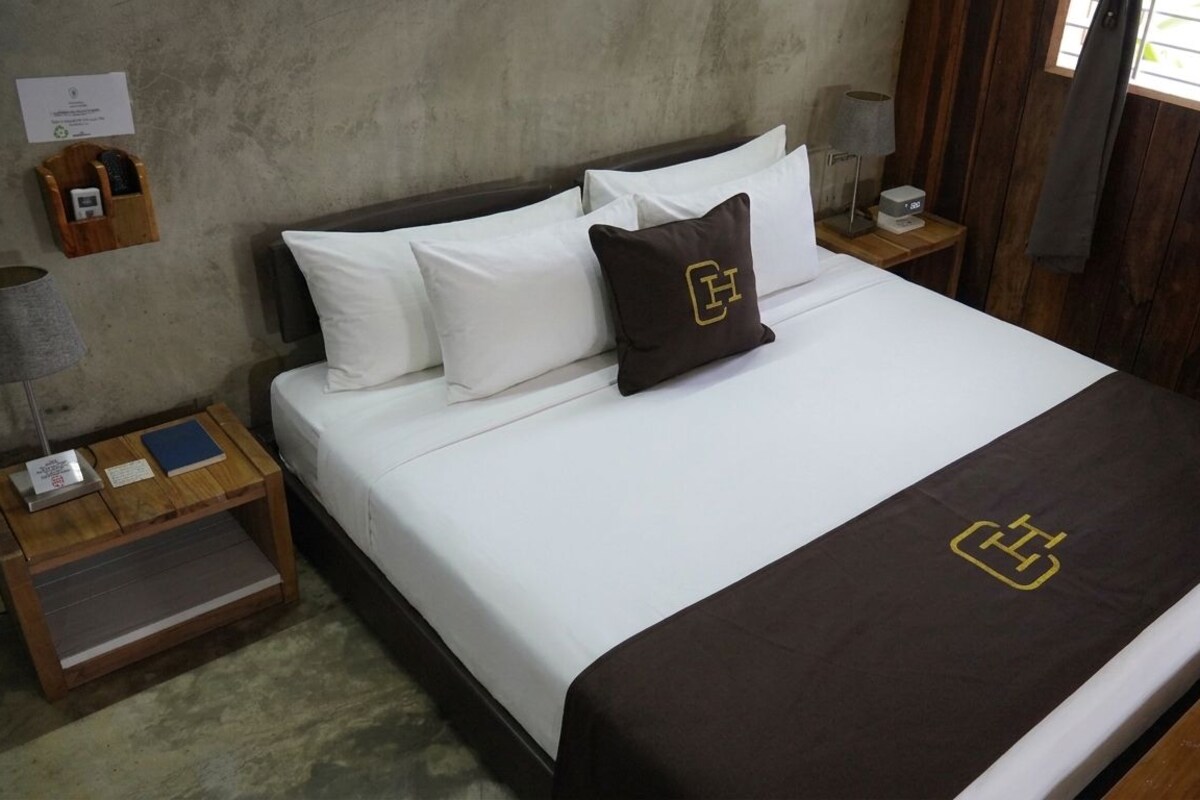 A neatly made bed with white and brown bedding, a wooden nightstand with a lamp and a small table, and a wooden wall.