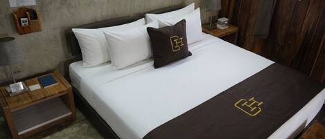 Cama bem arrumada com roupa de cama branca e castanha, mesa de cabeceira de madeira com abajur e uma pequena mesa, e uma parede de madeira.