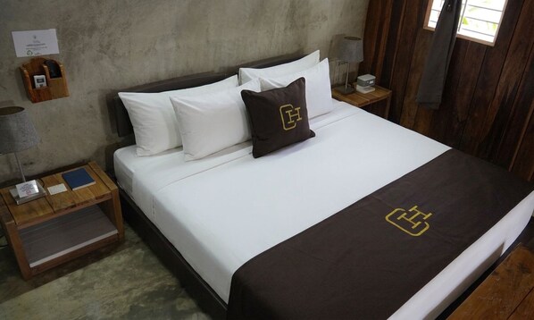 A neatly made bed with white and brown bedding, a wooden nightstand with a lamp and a small table, and a wooden wall.