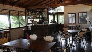 A spacious indoor area with a bar, wooden tables, wicker chairs, and large windows offering a view of greenery outside. - Hotel Central Bocas Del Toro (Panama)