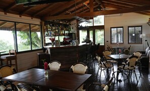 Un vaste espace intérieur comprenant un bar, des tables en bois, des chaises en osier et de grandes fenêtres donnant sur un paysage verdoyant.