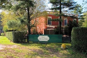 A hotel sign with the name 'Hotel De La Ville' in front of a red brick building surrounded by greenery. - Hotel De La Ville (Bereguardo)