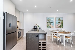 A modern kitchen with a built-in refrigerator, stove, and sink, and a dining area with white chairs and a wooden table. - Paradise Coast Luxe in the heart of Surfers Paradise (Surfers Paradise)