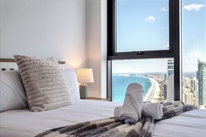Habitación de hotel con una cama, vista a la playa y al paisaje urbano a través de un ventanal grande, y una lámpara en la mesita de noche.