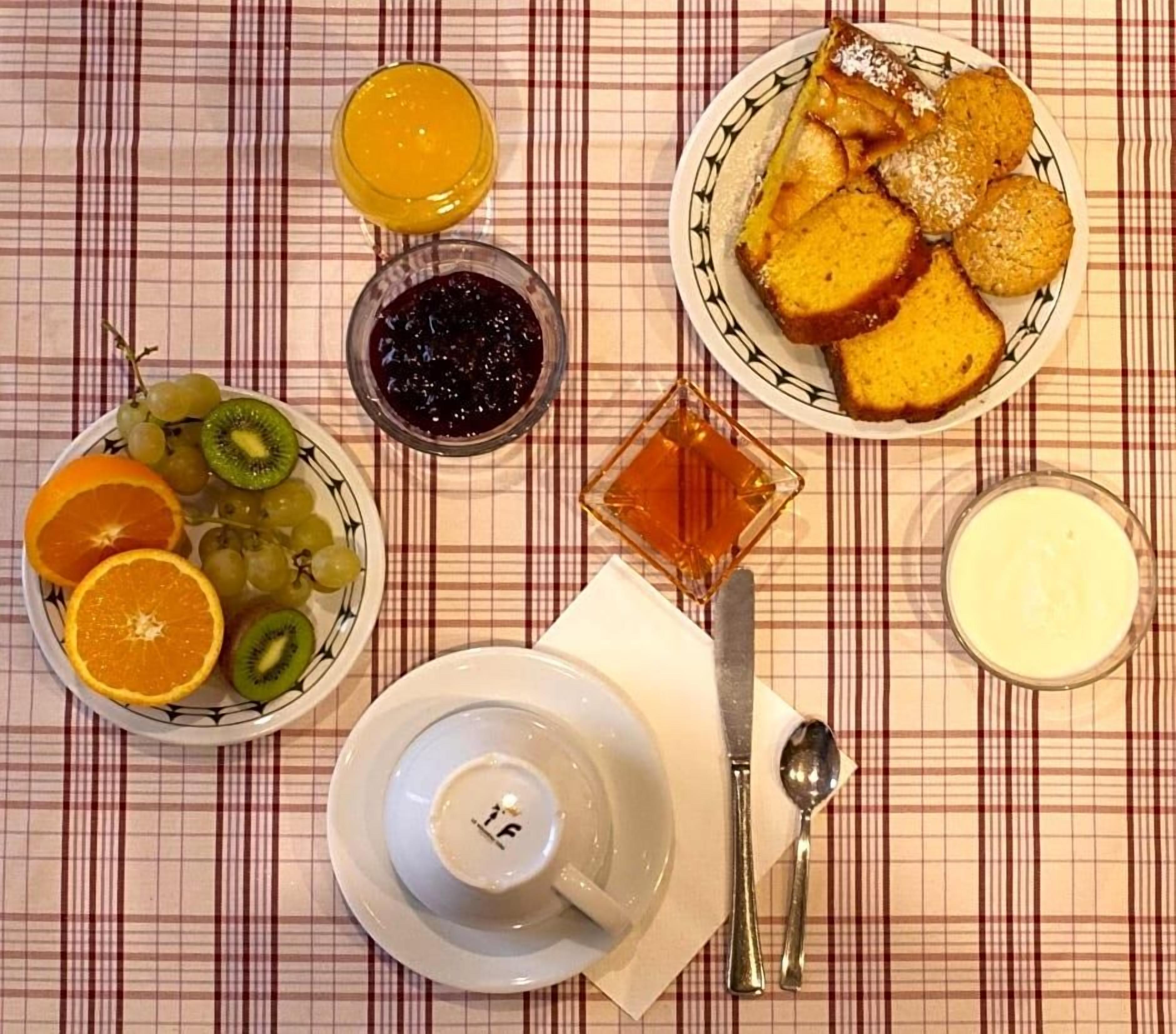 A table set with a plate of sliced bread, a bowl of fruit, a glass of orange juice, a bowl of jam, a small dish of honey, a cup of tea, and a glass of milk.