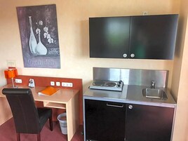 A compact kitchenette with a stove, sink, and cabinets, a desk with a chair, and a wall-mounted TV.