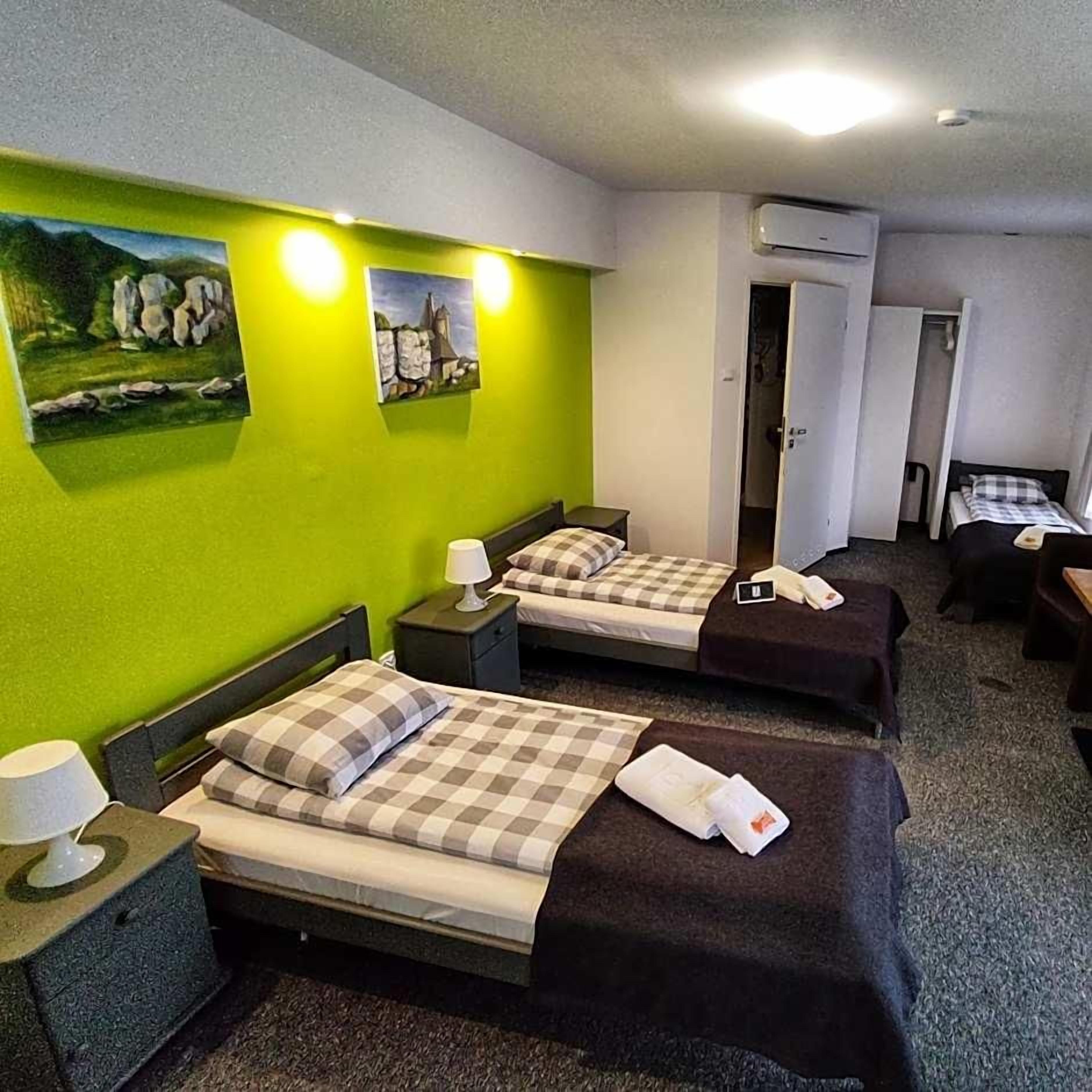 A hotel room with two beds, a desk, and a chair. There are paintings on the wall and an air conditioning unit.