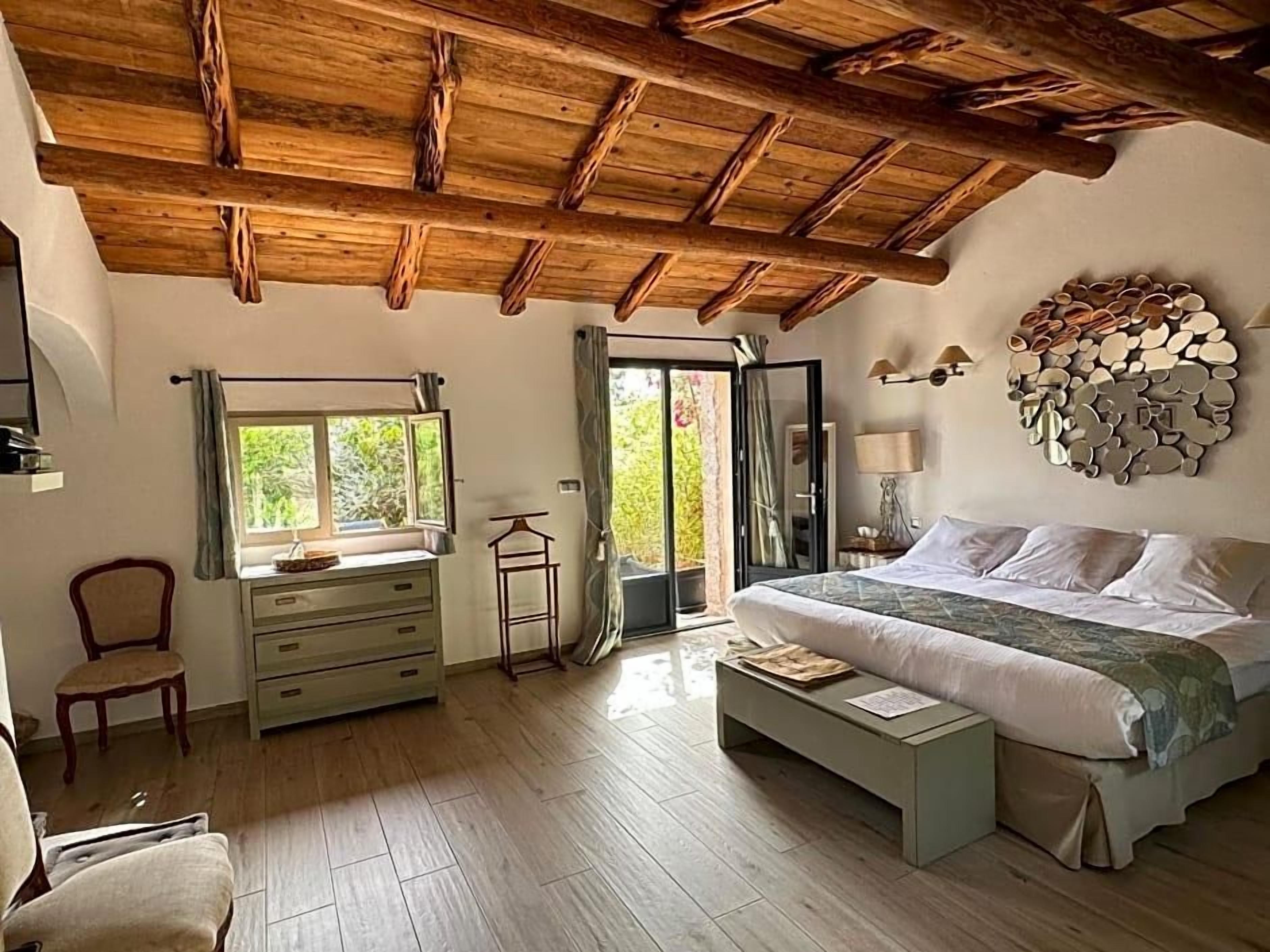 Une chambre à coucher avec un plafond en bois, un lit, une table de chevet, une chaise, une commode et une fenêtre avec des rideaux.