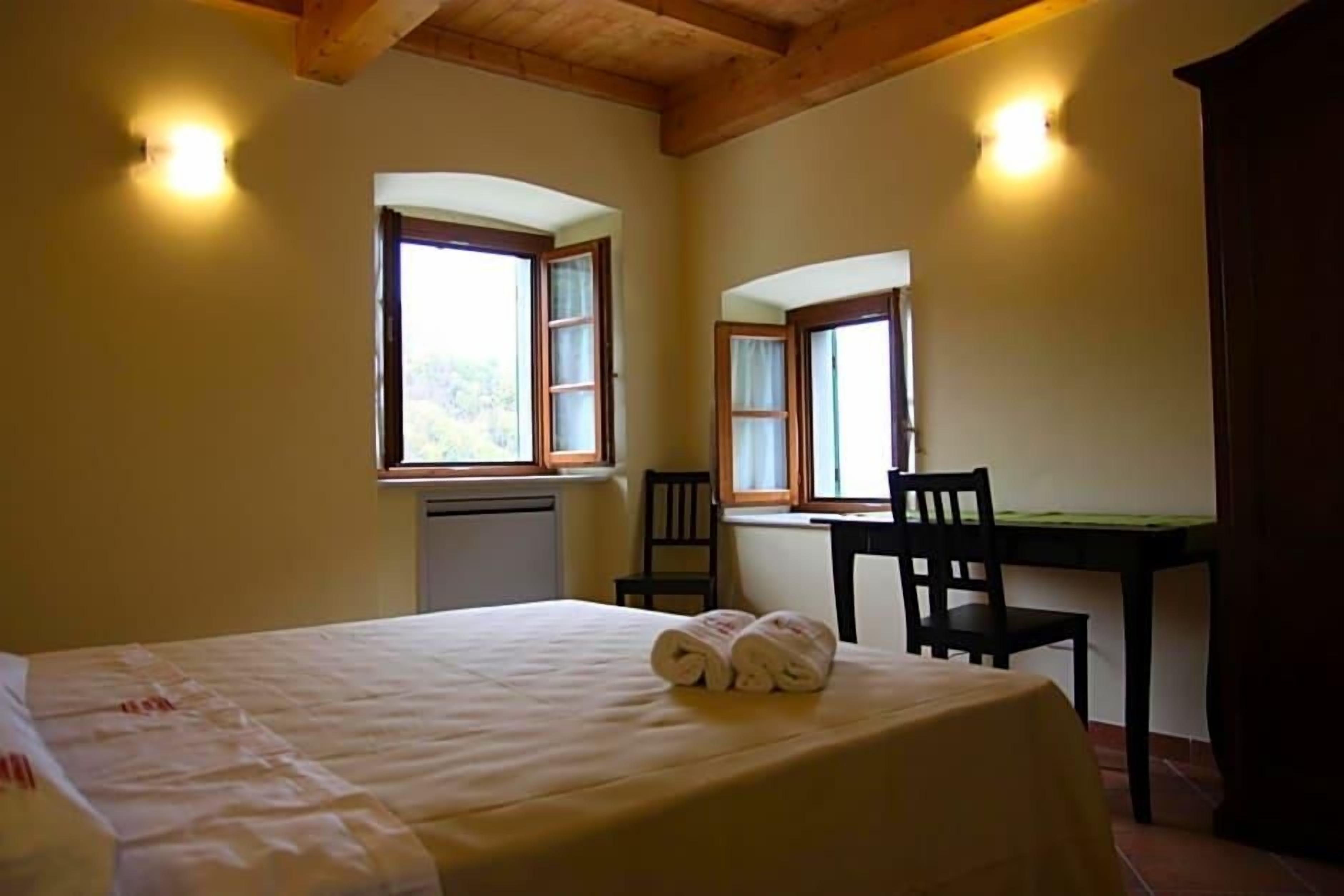 Una habitación con cama, dos sillones, una mesa y dos ventanas con cortinas.