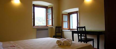 Una habitación con cama, dos sillones, una mesa y dos ventanas con cortinas.