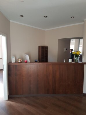 A reception area with a wooden reception desk, a small shelf with items, and a mirror reflecting a person and flowers. - Weltmann S Hotel Restaurant (Ennepetal)