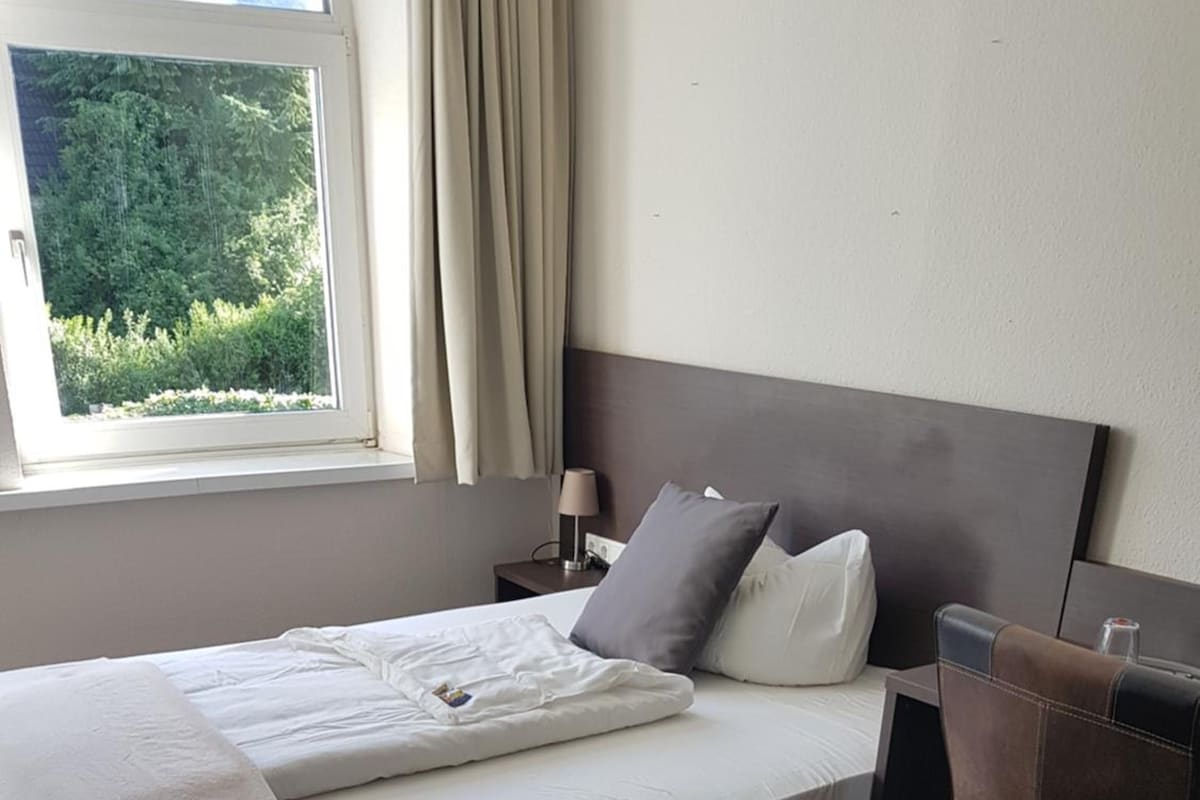 A neatly made bed with a white duvet and pillows, a bedside table with a lamp, and a view of greenery through a window.