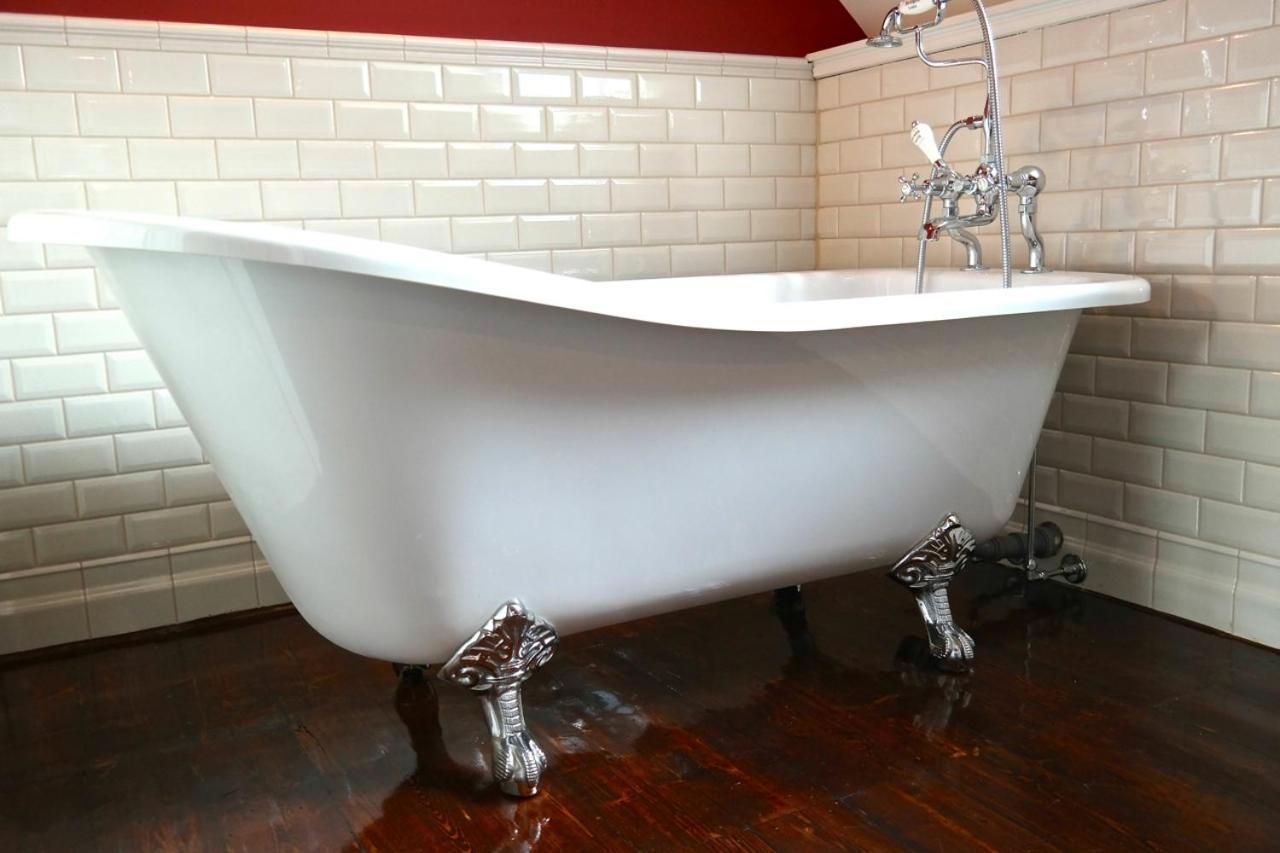 A freestanding bathtub in a corner with a white tiled wall and wooden floor.