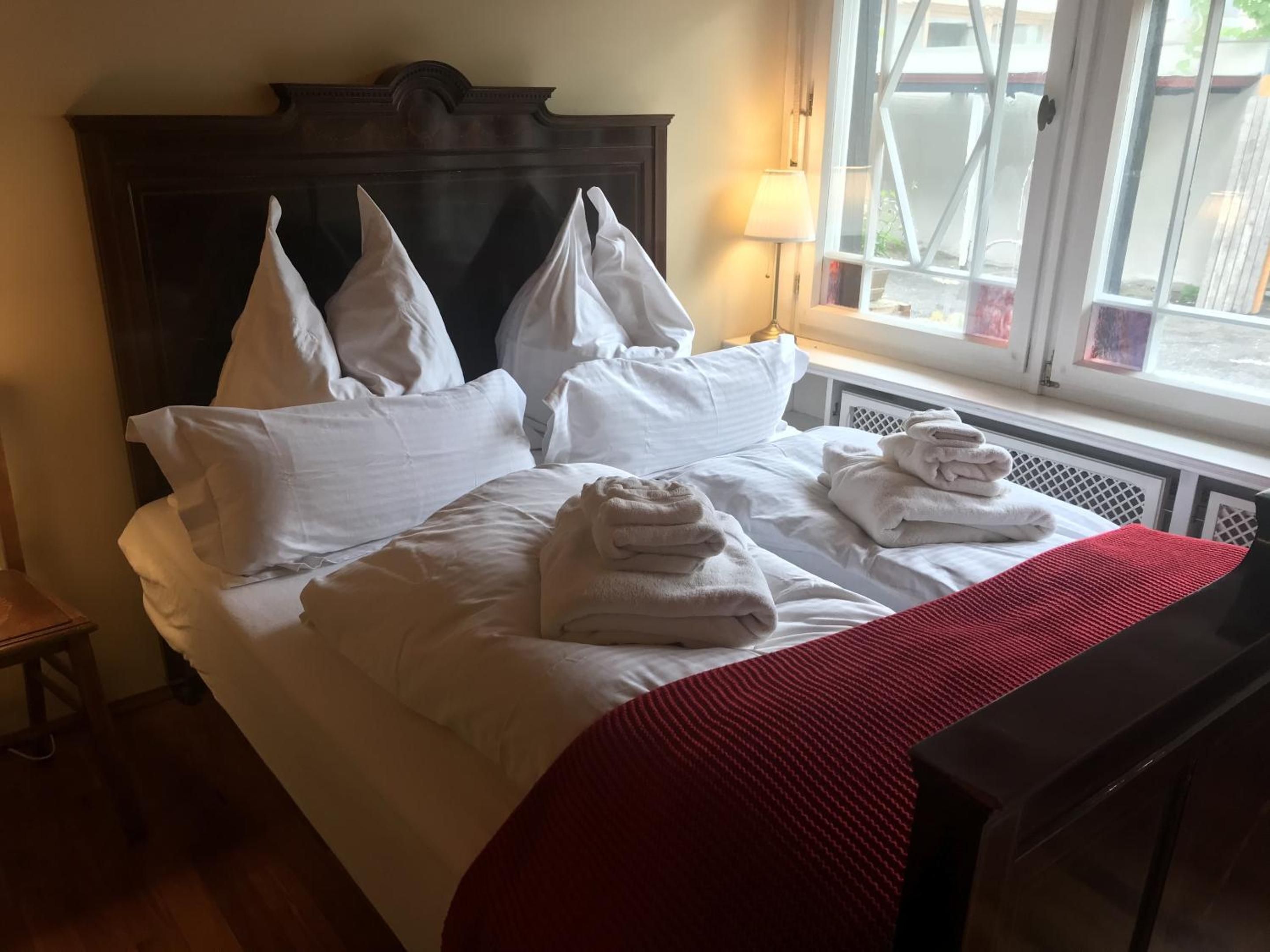 A hotel room with a large bed, white linens, a red throw blanket, and a window with a view of the outdoors.