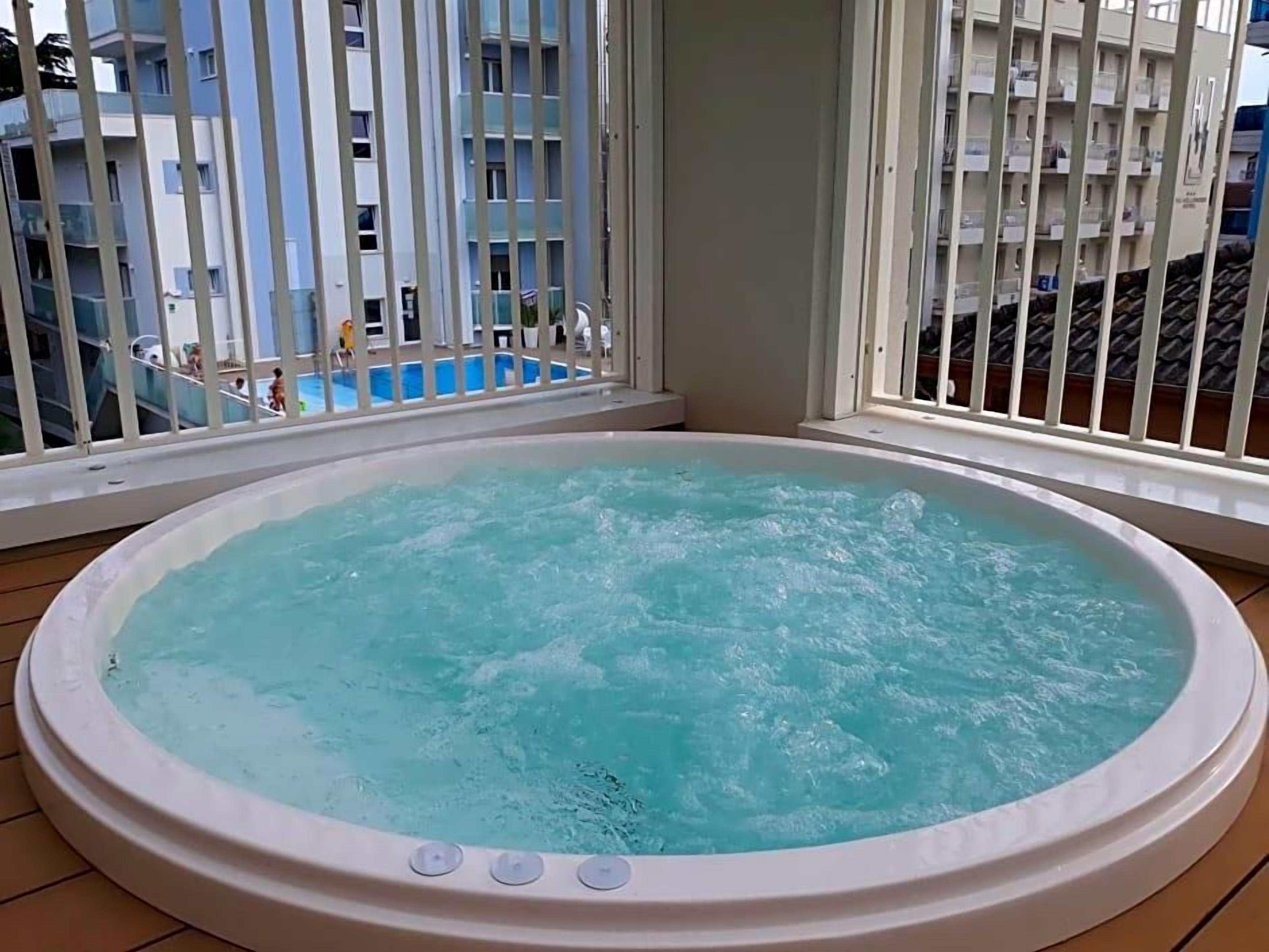 A hot tub on a balcony with a view of a swimming pool and a building in the background.