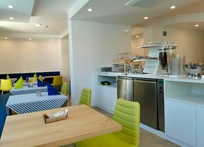 Un espace repas moderne avec une table en bois et des chaises vertes, une cuisine équipée d’appareils électroménagers en acier inoxydable et un plan de travail blanc sur lequel sont disposés divers objets.