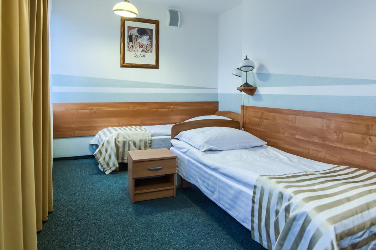 Una habitación con dos camas individuales, una mesita de noche, una lámpara y un cuadro en la pared.