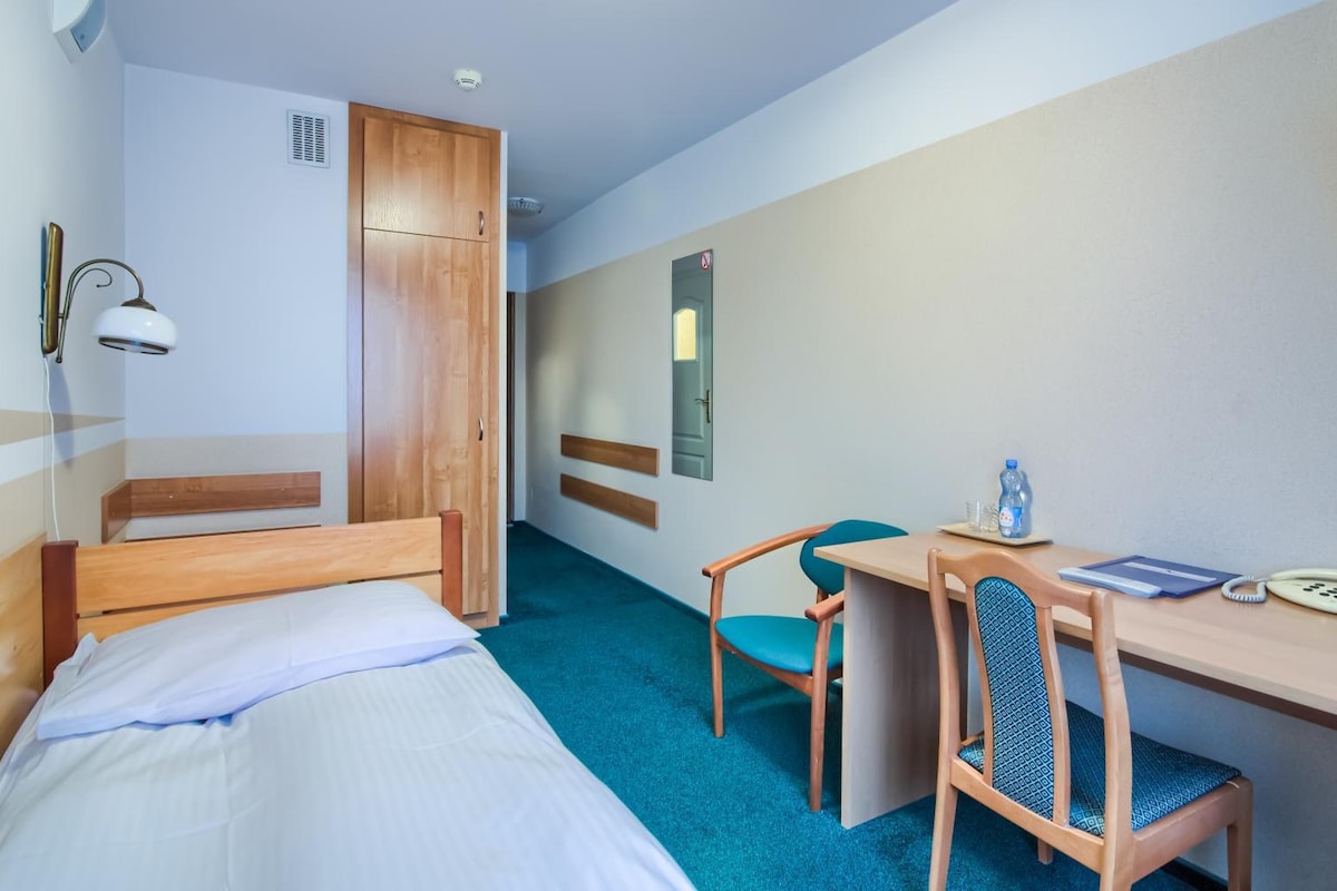 A hotel room with a bed, a desk with a chair, a water bottle, and a phone.