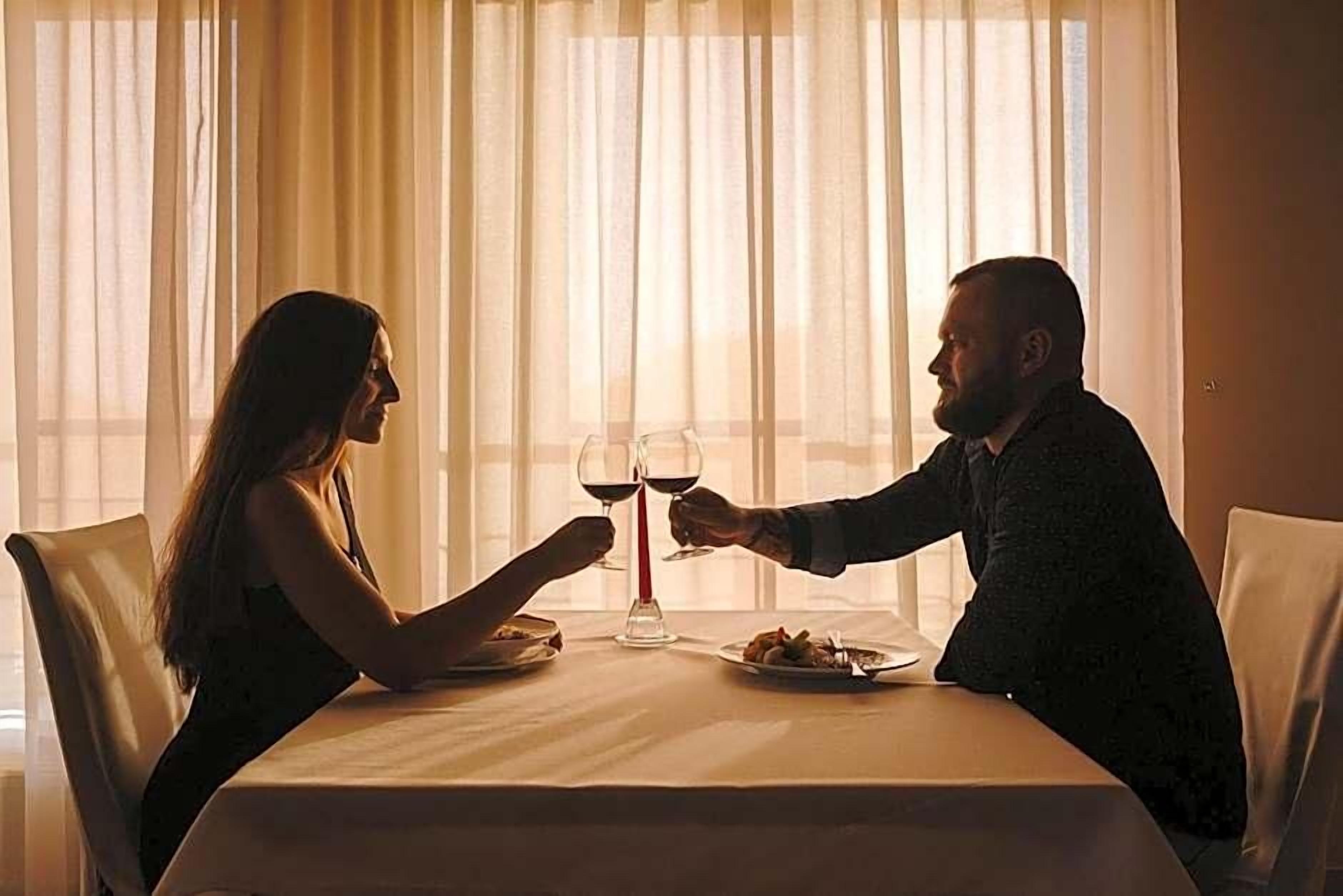 A couple seated at a table with wine glasses, a plate of food, and a window with sheer curtains in the background.