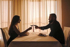 A couple seated at a table with wine glasses, a plate of food, and a window with sheer curtains in the background.