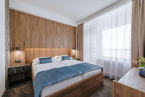 A modern hotel room with a wooden headboard, a large bed with a blue blanket, two bedside tables with lamps, and a view of the outdoors through sheer curtains.