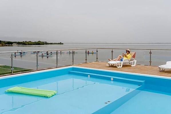 A swimming pool with a view of the sea, a lounge area with chairs, and a person relaxing.