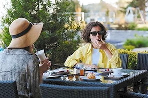 Two people seated at an outdoor table with a variety of food and drinks, surrounded by greenery.