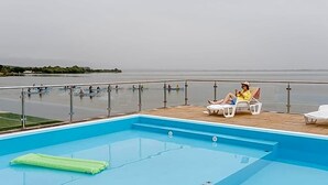 A swimming pool with a view of the sea, a lounge area with chairs, and a person relaxing.