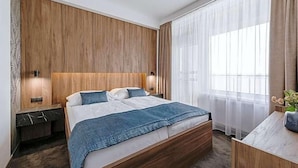 A modern hotel room with a wooden headboard, a large bed with a blue blanket, two bedside tables with lamps, and a view of the outdoors through sheer curtains.