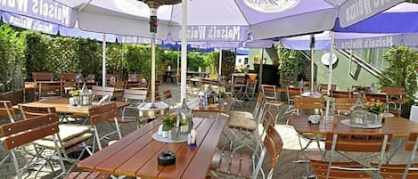 Un área de comedor al aire libre con mesas y sillas bajo sombrillas, rodeada de follaje.