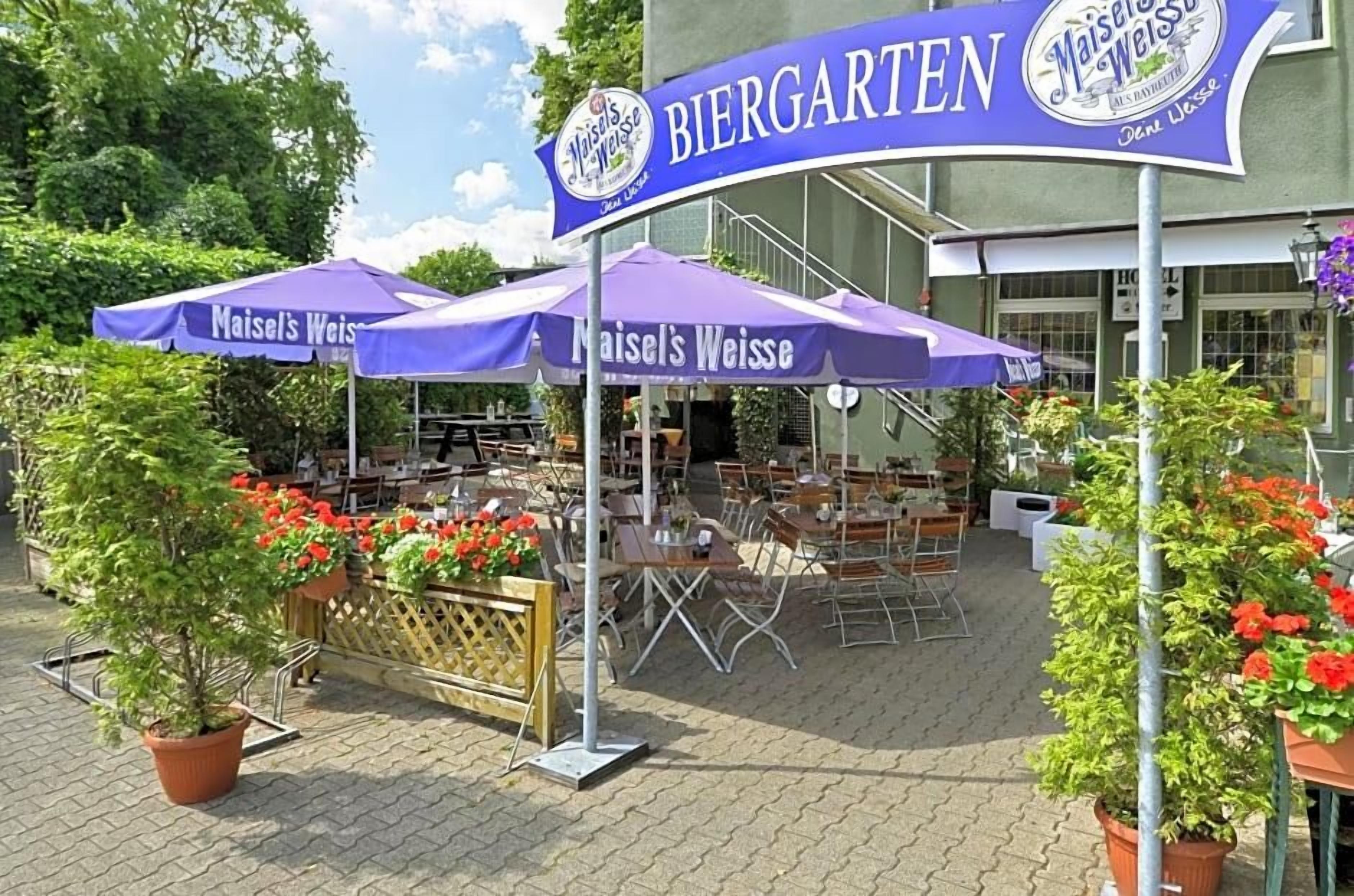 An outdoor seating area with tables and chairs under umbrellas, a sign for 'Maisel's Weisse Biergarten,' and potted plants with red flowers.