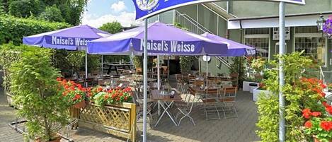 Un área de estar al aire libre con mesas y sillas bajo sombrillas, un letrero que dice 'Maisel's Weisse Biergarten' y plantas en macetas con flores rojas.
