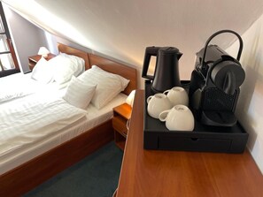 Una cama bien hecha con sábanas y almohadas blancas, una mesita de noche con una lámpara, y una cafetera con tazas sobre una bandeja.