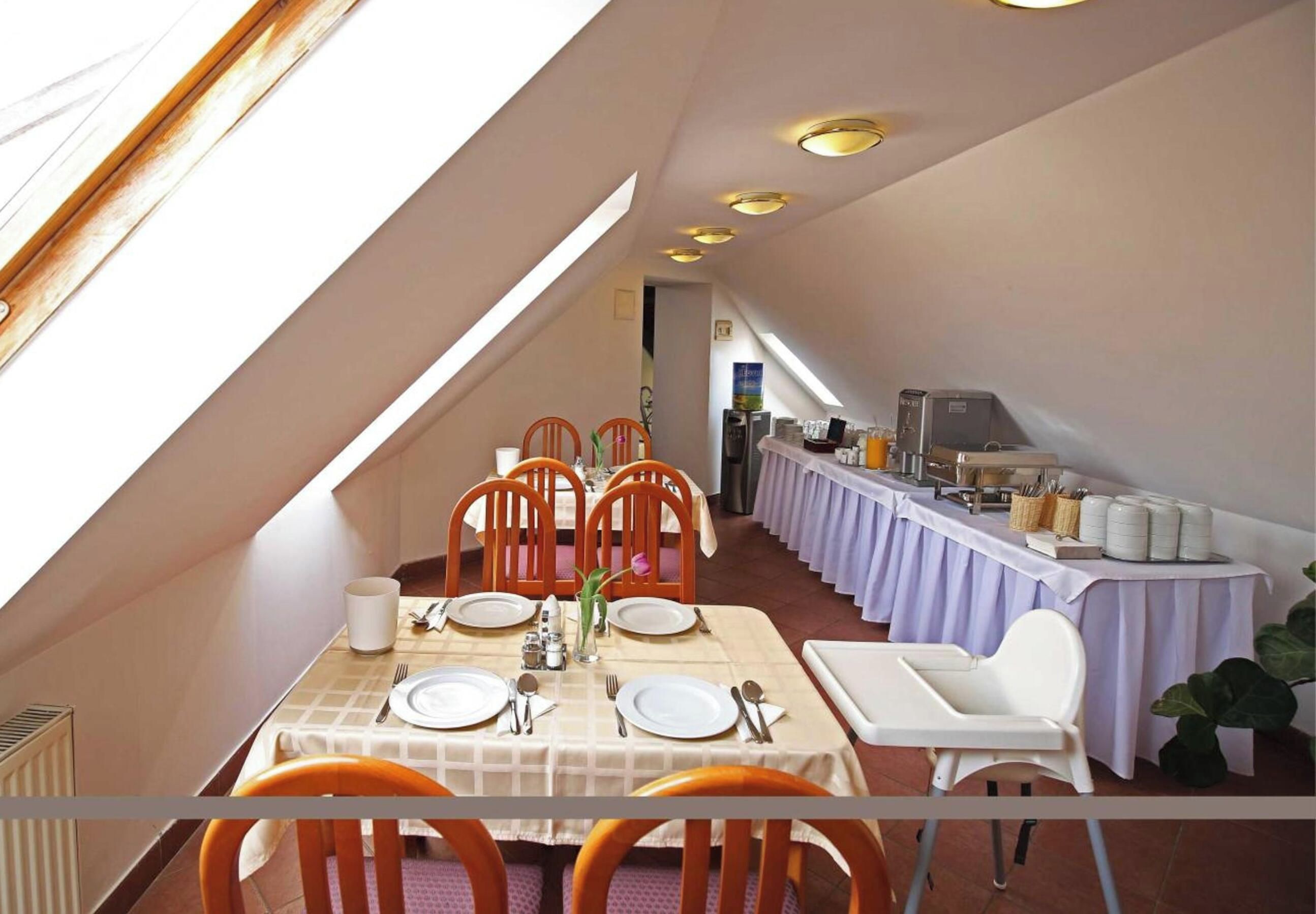 A dining area with a high ceiling and skylight, featuring a long table set for four with chairs, a buffet table with food and beverages, and a view of the adjacent room.