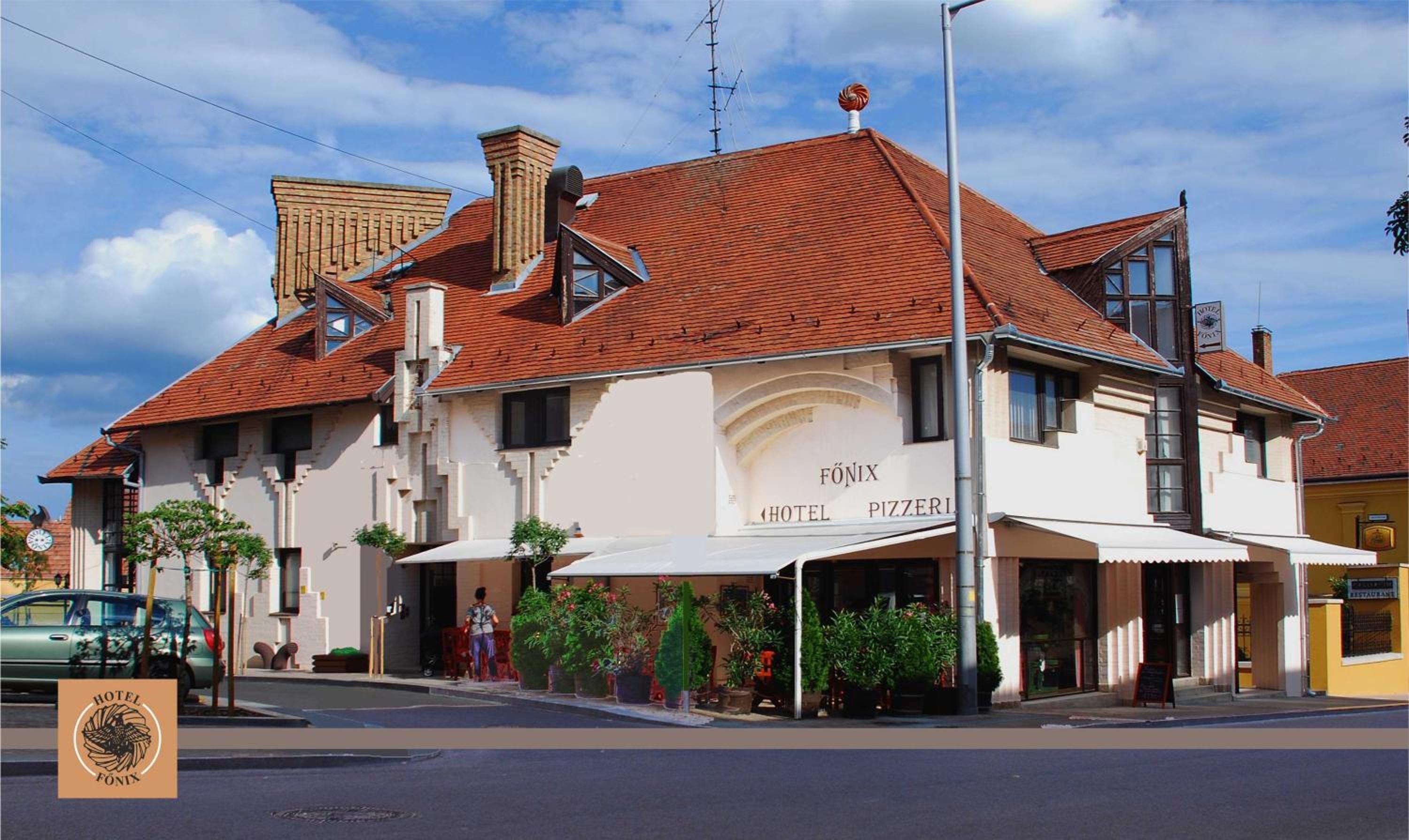 A hotel with a red-tiled roof, a sign for 'HOTEL FÖNIX PIZZERI', and outdoor seating with potted plants.