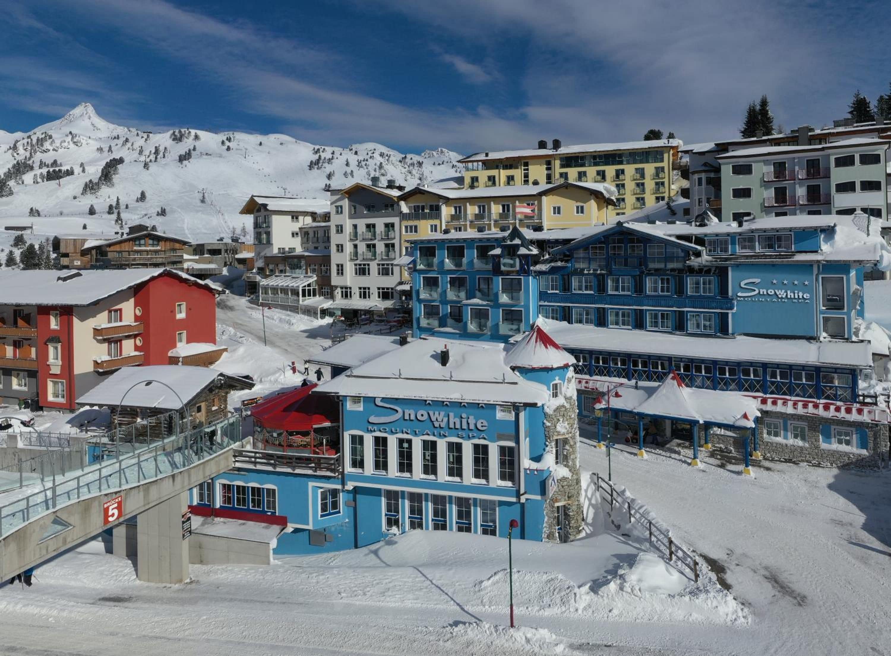 A snow-covered mountain town with a variety of buildings, including a prominent blue building with 'Snow White Mountain Spa' signage.