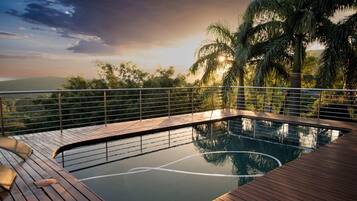A pool with a wooden deck and metal railing, surrounded by palm trees and a view of the landscape.
