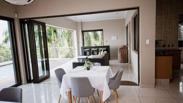 A dining area with a table set for four, a kitchen area, and a view of a balcony with greenery.