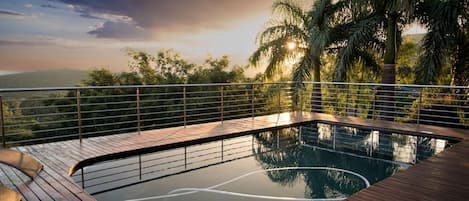 A pool with a wooden deck and metal railing, surrounded by palm trees and a view of the landscape.
