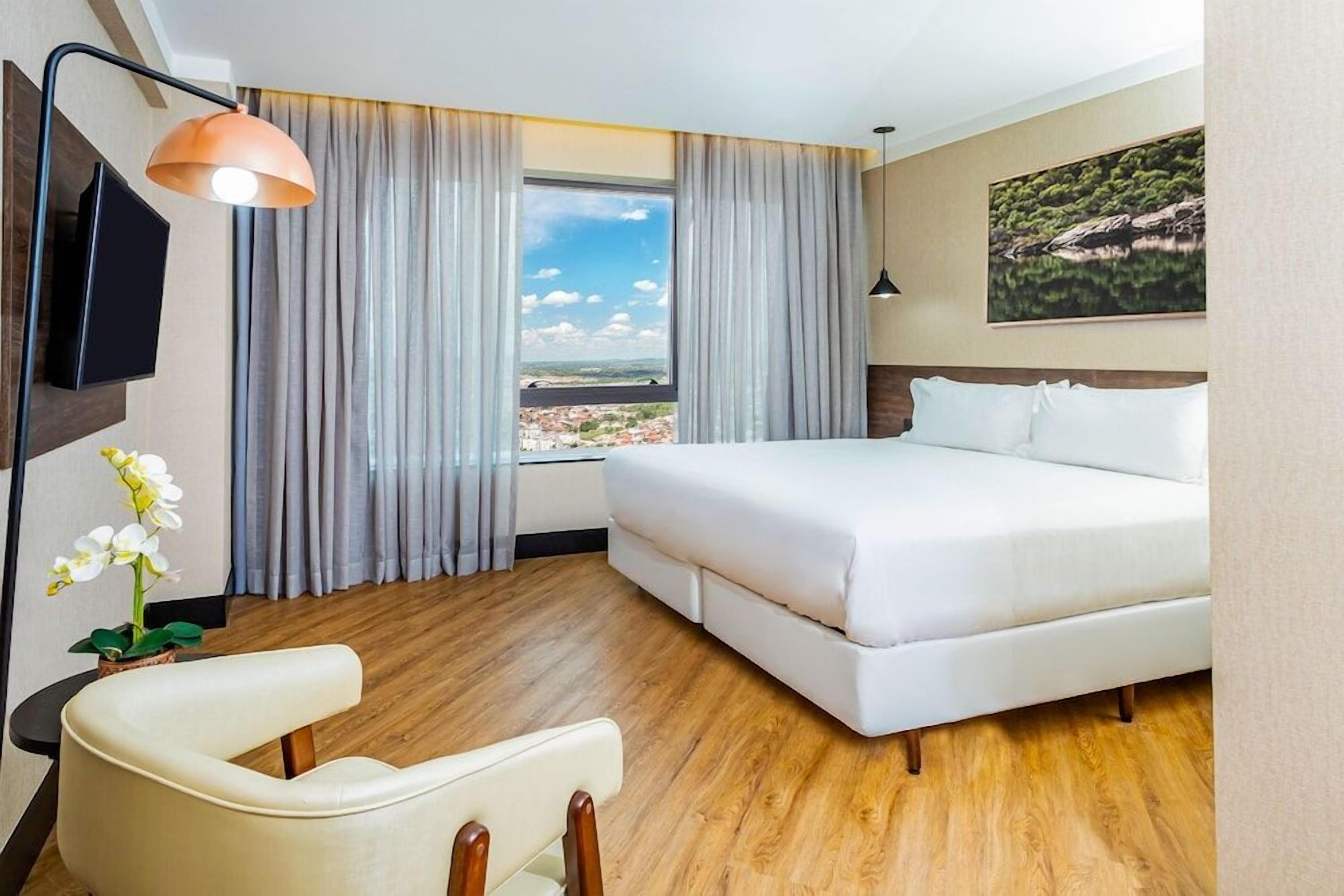 Una habitación de hotel con cama, escritorio, silla, televisor, ventana con cortinas y un cuadro en la pared.