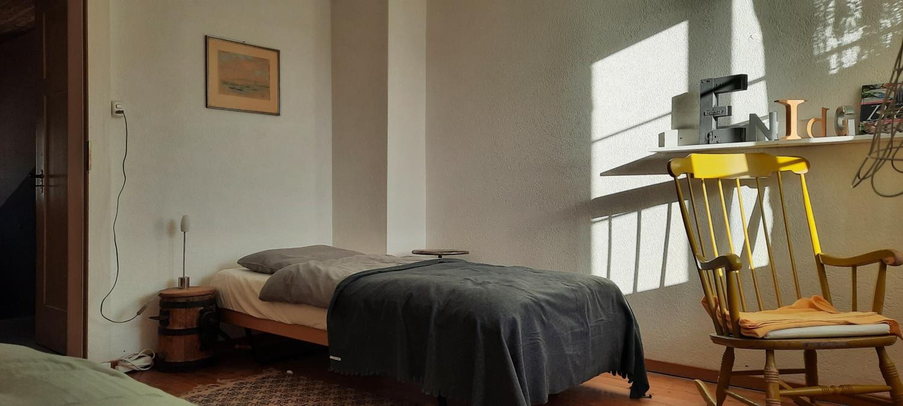Ein Schlafzimmer mit einem Bett, einem Sessel, einem Schreibtisch mit einem Computer und einem Fenster.