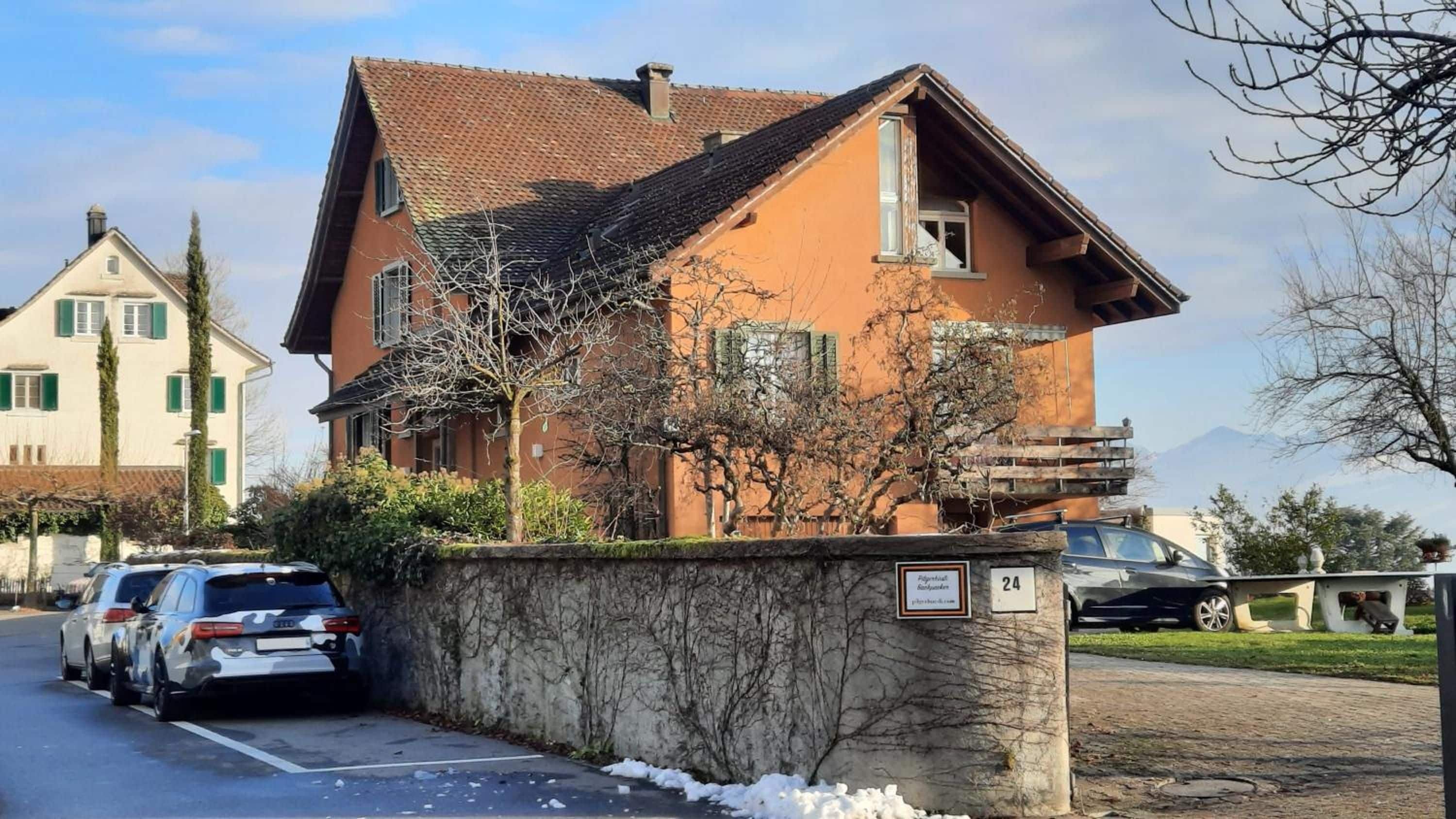 A two-story building with a sloped roof, a stone wall in front, and parked cars.